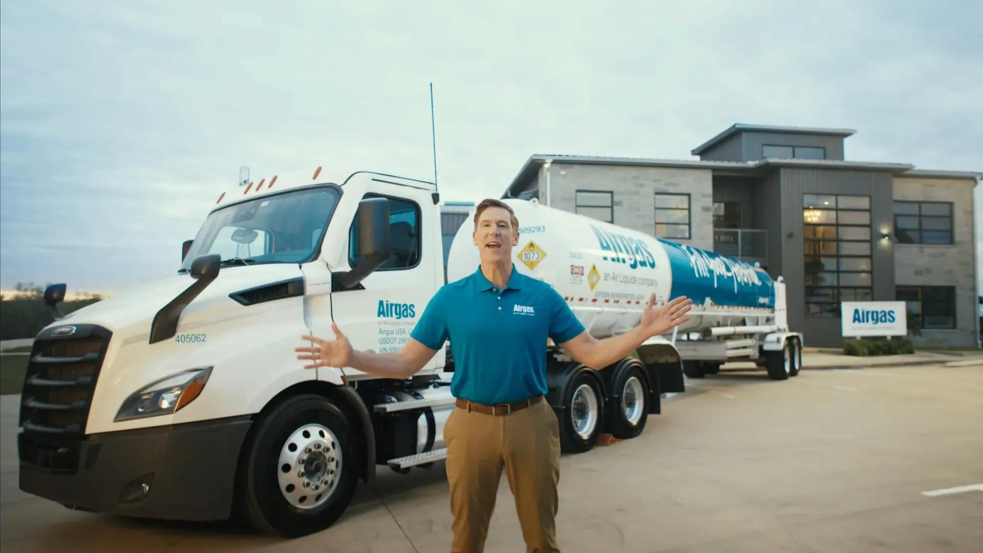 A man stands smiling in front of a large white and blue Airgas truck, arms spread wide. The scene is set in an industrial area with a modern building in the background. The atmosphere is professional and welcoming.
